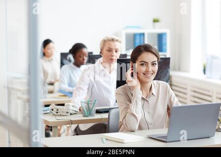 Portrait of female helpdesk operators sitting at in row , focus on smiling woman looking at camera while talking to customer via headset, copy space Stock Photo
