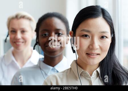 Multi-ethnic group of female call center operators looking at camera standing in row, focus on smiling Asian woman wearing headset in foreground, copy Stock Photo