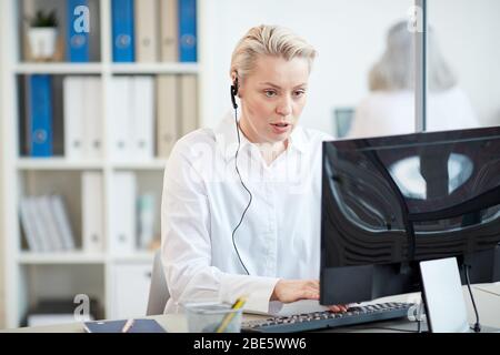 Portrait of female manager wearing headset and using computer while answering customer service request in office interior, copy space Stock Photo