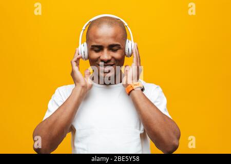 Portrait of an attractive smiling confident casual young african man standing over yellow background, listening to music with wireless headphones Stock Photo