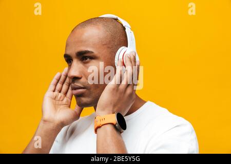 Portrait of an attractive confident casual young african man standing over yellow background, listening to music with wireless headphones Stock Photo