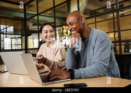 Image of multiethnic young female and male coworkers sitting at table and working on laptops in office Stock Photo