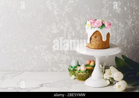 Easter cake with creamy roses on white stand. White flowers with holiday cupcakes. Cement wall. Copy space Stock Photo