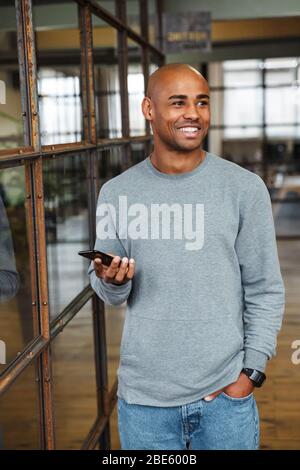 Image of young attractive bald african american man wearing sweatshirt holding cellphone while working in office Stock Photo