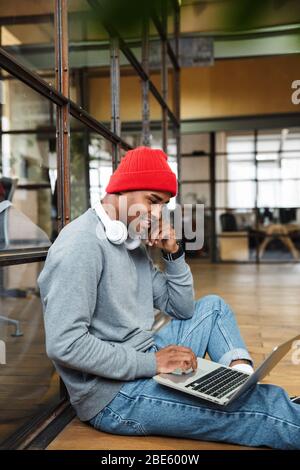 Image of young attractive african american man wearing hat using laptop computer while working in office Stock Photo