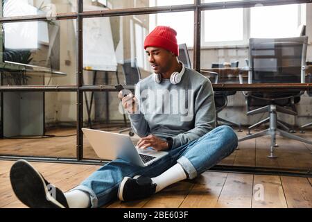 Image of a concentrated african young man sit indoors on floor using laptop computer and mobile phone. Stock Photo