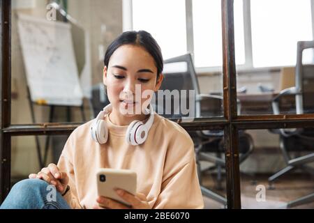 Image of young beautiful asian woman smiling and holding cellphone while working in office
