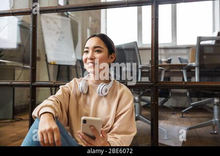 Image of young beautiful asian woman smiling and holding cellphone while working in office Stock Photo