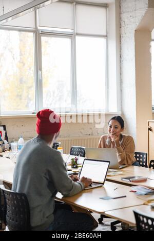 Image of a young concentrated multiracial colleagues indoors work in office with laptop computer talking with each other. Stock Photo