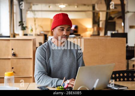 Image of young attractive african american man wearing hat using laptop computer while working in office Stock Photo