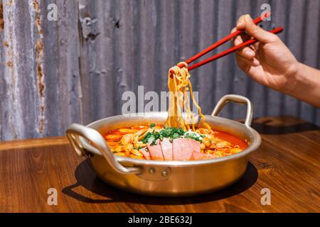 Hand pinching noodles with chopsticks. Traditional Korean ramen soup with kimchi, ham, sausage and cheese in silver bowl on wooden table. Stock Photo