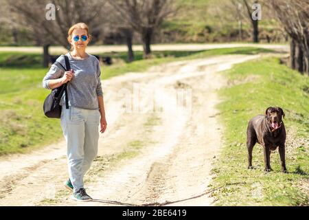 Woman in sunglasses staying on a road, with dog chocolate Labrador retriever. Stock Photo