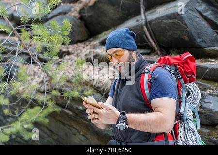 A man tourist with a backpack stands near a sheer stone rock and myotrites in the screen of a mobile phone. Searches for a way using GPS navigator. Th Stock Photo
