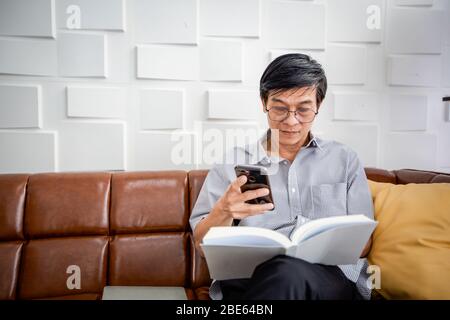 Senior Asian man reading book and using smart phone on sofa in living room at home  Portrait of Asian elderly man is Relaxing and Happiness With Read Stock Photo