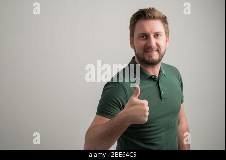 Portrait of serious stylish attractive man with thick beard, dressed in casual green t shirt showing like gesture isolated on gray background with cop Stock Photo