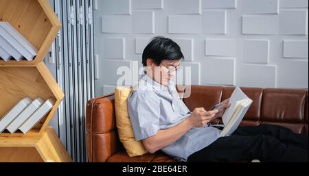 Senior Asian man reading book and using smart phone on sofa in living room at home  Portrait of Asian elderly man is Relaxing and Happiness With Read Stock Photo