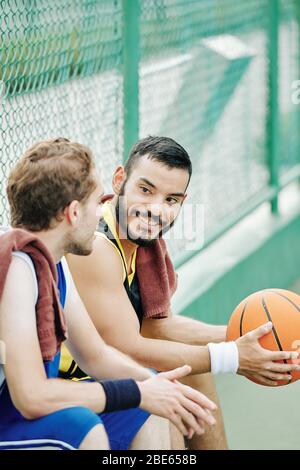 Smiling handsome young Hispanic man with basketball ball looking at friend telling funny story Stock Photo