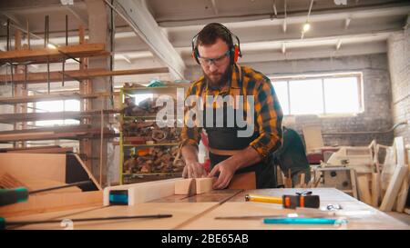 Bearded man cutting the wood using a circular saw in the workshop. Mid shot Stock Photo