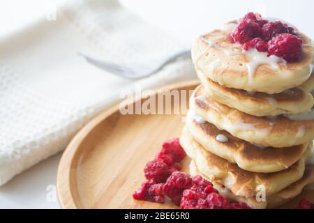 Fluffy pancakes with condensed milk and raspberries on a wooden plate with a kitchen napkin and a fork Stock Photo