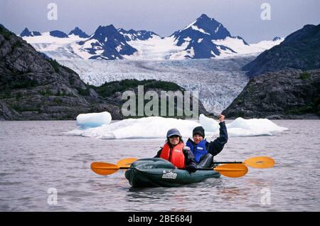 Mother and son in warm jackets, hats and life vests paddle a kayak past icebergs in Halibut Cove, Homer, Alaska, USA on a summer day. Stock Photo