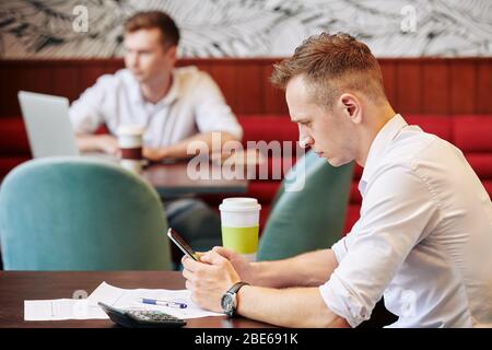Serious frowning young businessman sitting at cafe table with documents in front of him and reading news on smartphone screen Stock Photo