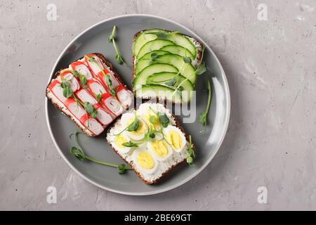 Three sandwiches on toast with cream cheese, cucumbers, quail eggs and crab sticks decorated with microgreens peas on a plate on gray concrete Stock Photo
