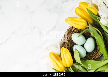 Beautiful bouquet of yellow tulips on marble table. Three blue painted eggs in rustic nest. Happy Easter concept. Top view. Copy space Stock Photo