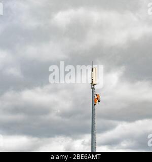 Technician working/ climbing up a mobile phone mast against grey cloudy sky. Service provider unknown. Stock Photo