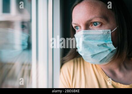 Woman in self-isolation during virus outbreak looking through window. Worried female person with protective surgical mask in stay at home concept, sel Stock Photo