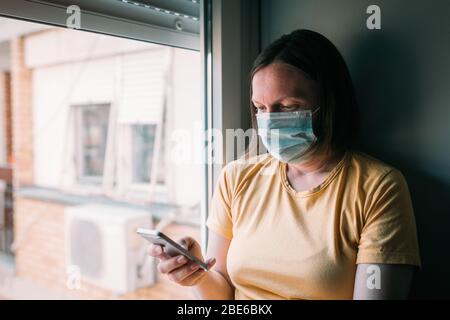 Woman in self-isolation during virus outbreak using mobile phone. Worried female person with protective surgical mask, holding smartphone in stay at h