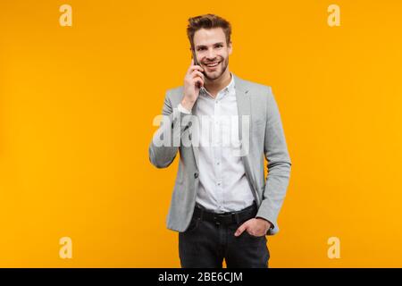 Handsome smiling young businessman wearing jacket standing isolated over yellow background, talking on mobile phone Stock Photo