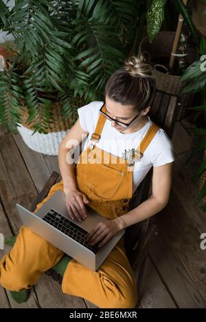 Young female gardener in glasses wearing overalls, sitting on wooden chair in greenhouse, using laptop after work, communicates on internet with custo Stock Photo