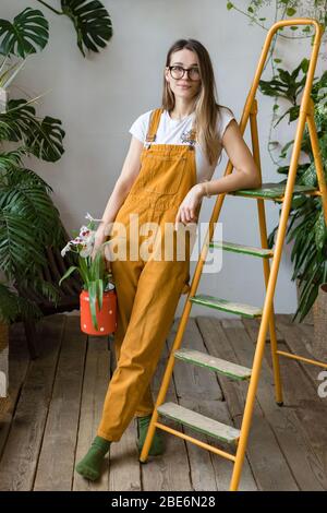 Young woman gardener in glasses wearing overalls, holding orchid in old red milk can and leaning on orange vintage ladder, looking at camera. Home gar Stock Photo
