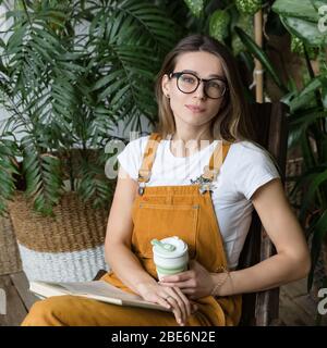 Young female gardener in glasses wearing overalls, resting, sitting on wooden chair in home greenhouse, hold collapsible silicone eco muge coffee/tea Stock Photo