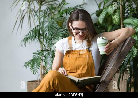 Young female gardener in glasses wearing overalls, resting after work, sitting on wooden chair in home greenhouse, holding reusable foldable coffee/te Stock Photo