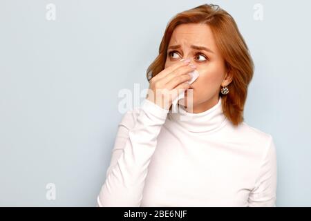 Studio portrait of scared young woman in white turtleneck with napkin blowing nose, looking to the source of allergy, place for advertising. Rhinitis, Stock Photo
