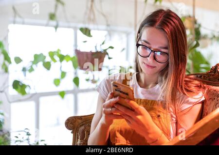 Young female gardener in glasses wearing overalls, sitting on vintage chair in greenhouse and using smartphone, typing message, selective focus. Plant Stock Photo