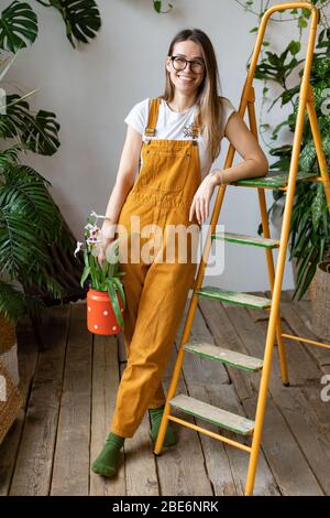 Young woman gardener in glasses wearing overalls, holding orchid in old red milk can and leaning on orange vintage ladder, smiling and looking at came Stock Photo