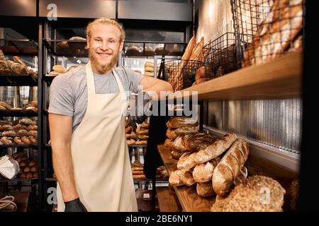 Small Business.Bearded man assistant standing at bakery shop leaning on shelf with bread posing to camera smiling cheerful Stock Photo