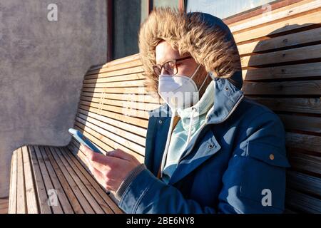 Sad man with hood sitting alone on bench in sunny weather, wearing facial mask against transmissible infectious diseases, coronavirus pandemic, using Stock Photo