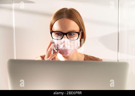 Woman freelancer in glasses wearing medical protective mask, talking on smartphone, online working from home during self isolation and quarantine. Wor Stock Photo