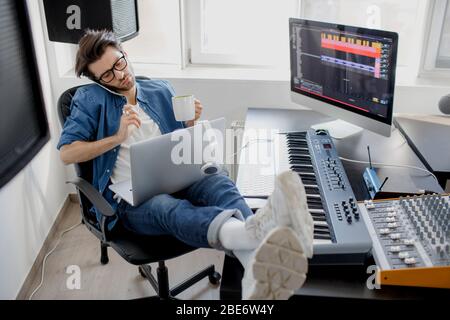 Talking on the phone and working on a laptop. Busy guy. Multitasking concept. DJ in broadcasting studio. music producer is composing a song on Stock Photo