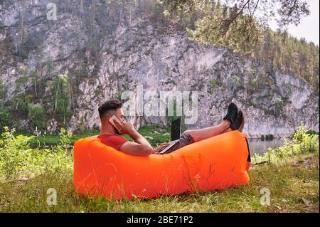 Bearded man is working and smiling with laptop at the park under the tree. Happy freelancer is sitting and using app or website on the grass. Work Stock Photo