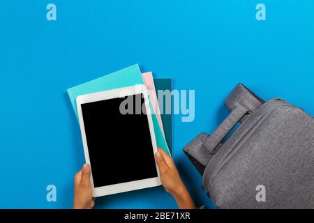 Kid hands with tablet computer, notebooks, books and gray backpack over light sky blue background Stock Photo