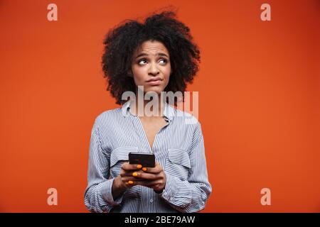 Confused young african woman using mobile phone isolated over red background Stock Photo