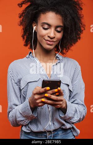 Smiling young beautiful african woman wearing casual clothes standing isolated over red background, listening to music with earphones and mobile phone Stock Photo