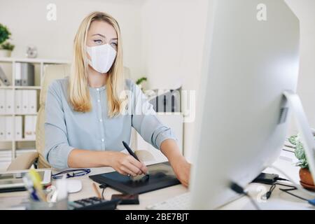 Young female graphic designer with medical mask on her face using graphics tablet while working with computer at studio or office. Stock Photo