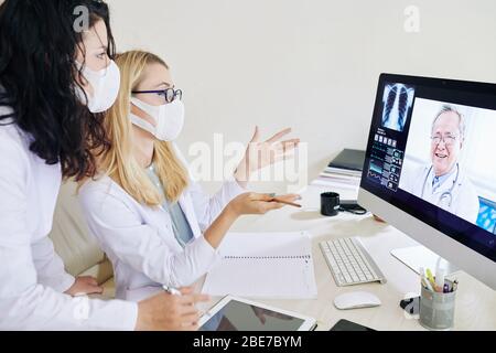 Young doctors in medical masks having video conference with experienced pulmonologist and discussing symptoms of coronavirus Stock Photo