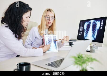 Professional young female pulmonologists pointing at lungs x-ray image on screen of tablet computer when having video conference with colleagues Stock Photo