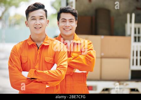Smiling Asian moving service workers standing in front of truck loaded with cardboard boxes Stock Photo